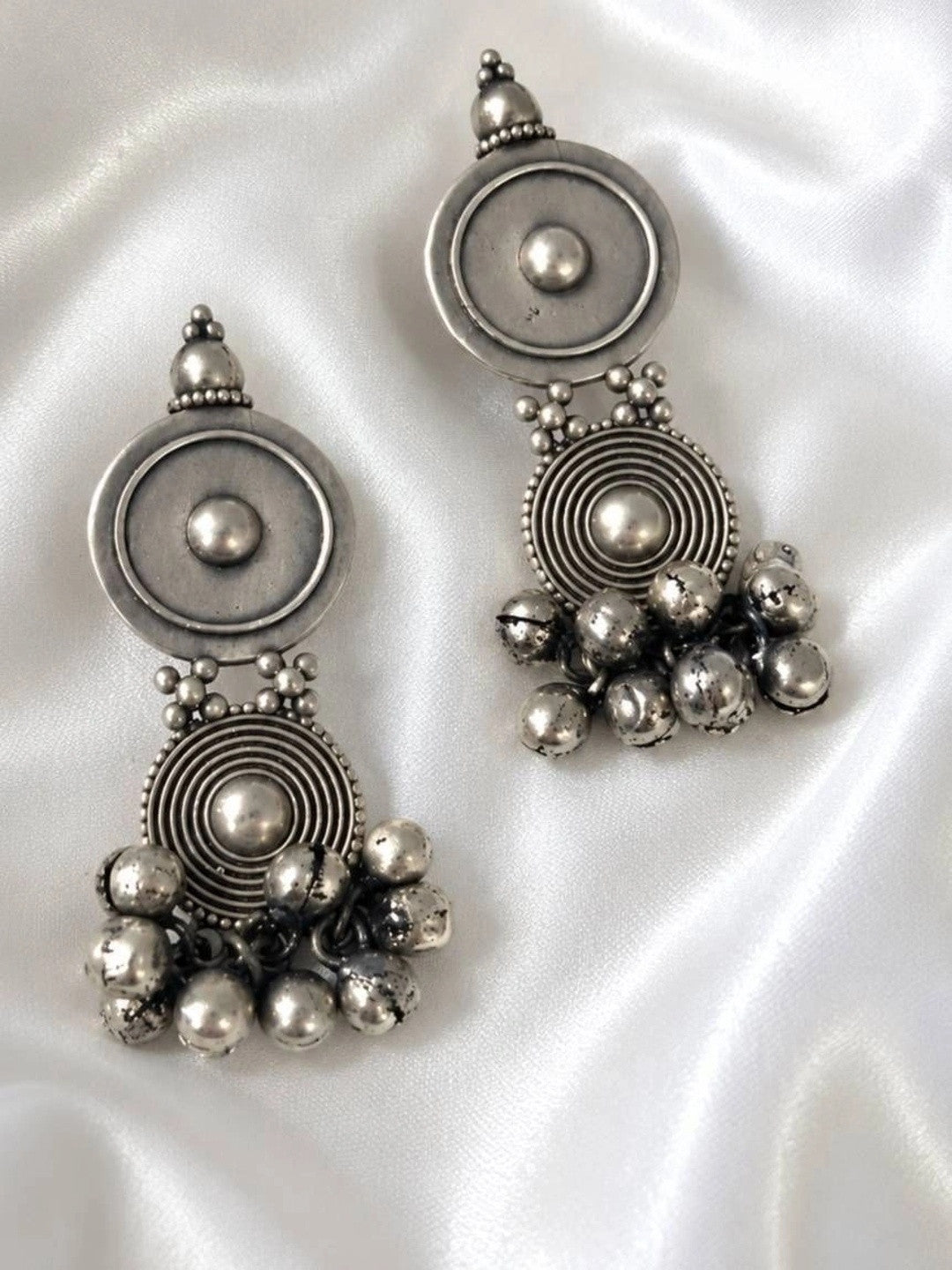 Side by side view of oxidised silver dual disc ghungroo earrings on white satin fabric