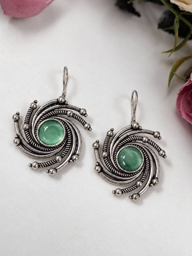 Front view of oxidised silver swirl earrings with green stone centres laid flat on a white surface beside pink roses