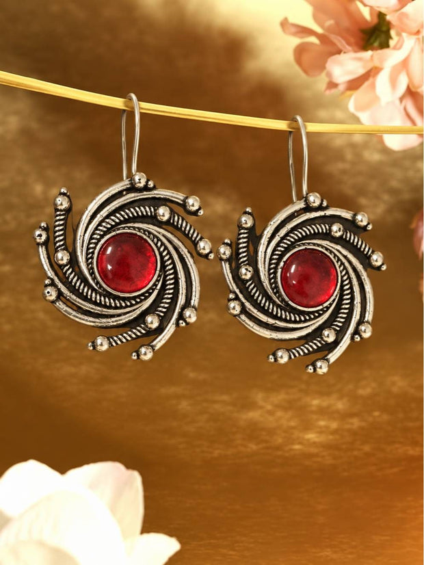 Pair of oxidised silver spiral drop earrings with red cabochon centre stones hanging from a gold rod against a warm golden background