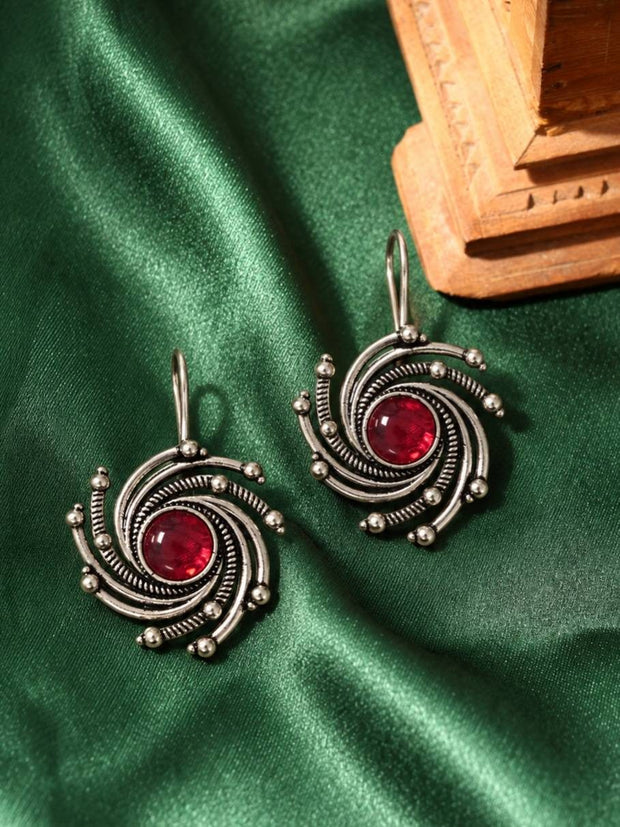 Two oxidised silver swirl earrings with red glass stones laid flat on green satin fabric beside a carved wooden block