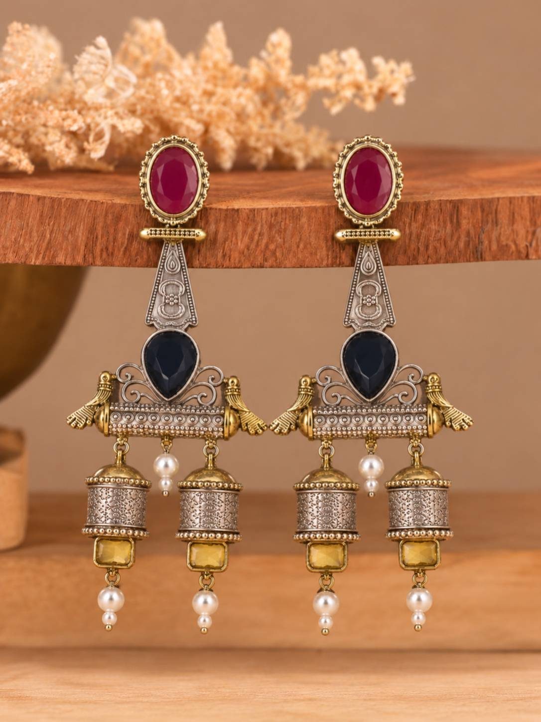 Pair of oxidised dual-tone jhumka earrings displayed upright on a wooden prop showing crimson, navy and amber stones with pearl drops