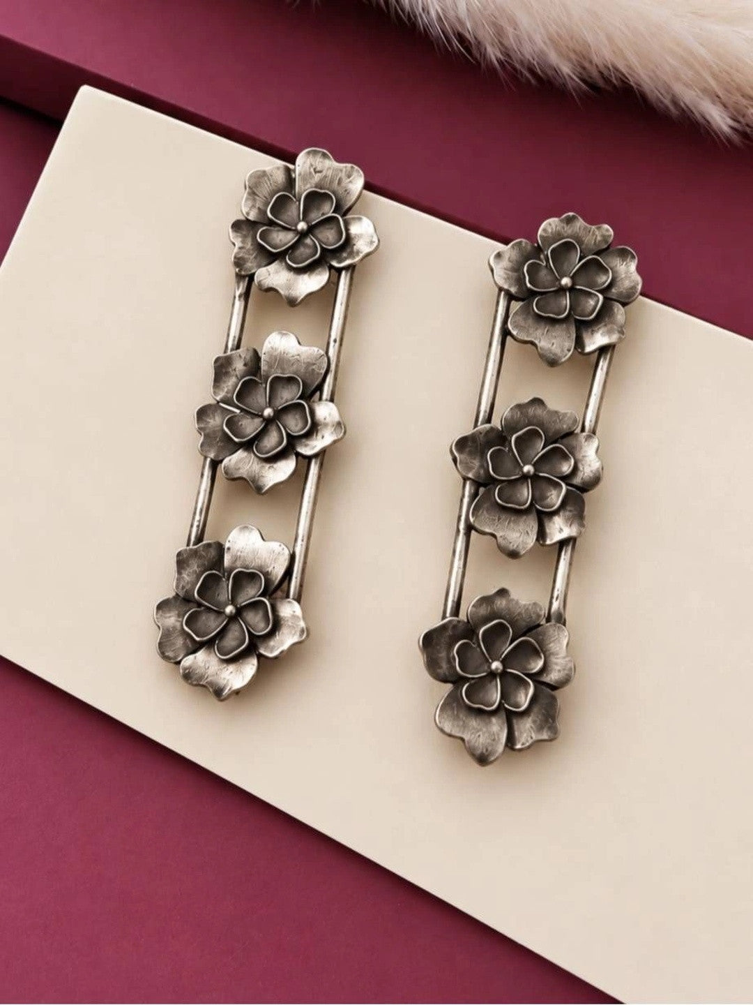 Pair of oxidised silver triple floral bar drop earrings displayed on a cream surface with burgundy backdrop