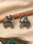 Pair of oxidised silver camel stud earrings laid flat on cream fabric beside a wooden stick and peacock feather