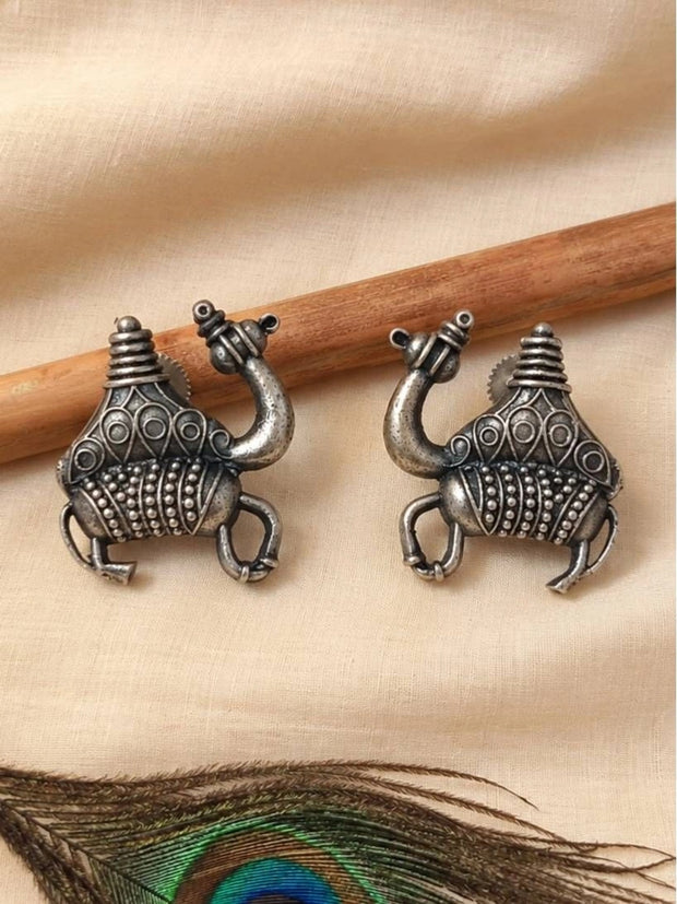 Pair of oxidised silver camel stud earrings laid flat on cream fabric beside a wooden stick and peacock feather