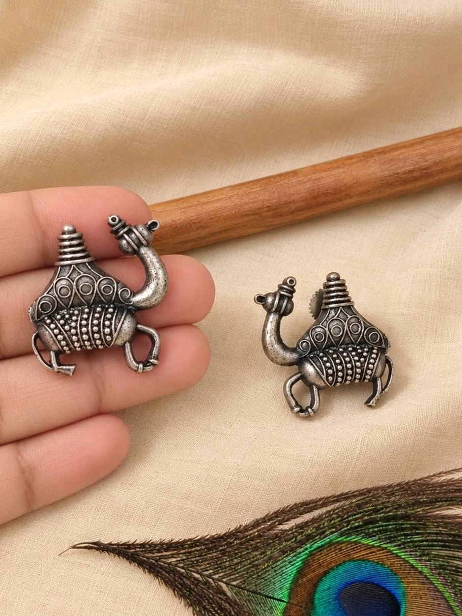 Hand holding one oxidised silver camel earring beside its pair resting on cream fabric with peacock feather