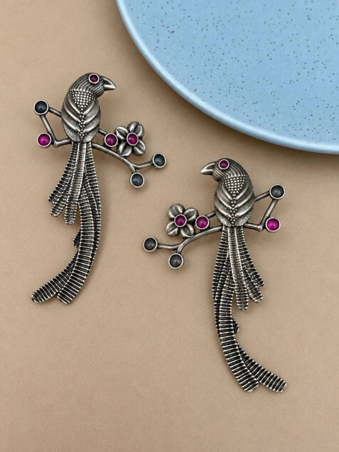 Pair of oxidised silver bird branch drop earrings with pink and grey stone accents on beige surface