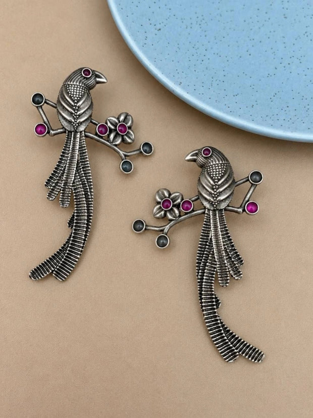 Pair of oxidised silver bird branch drop earrings with pink and grey stone accents on beige surface