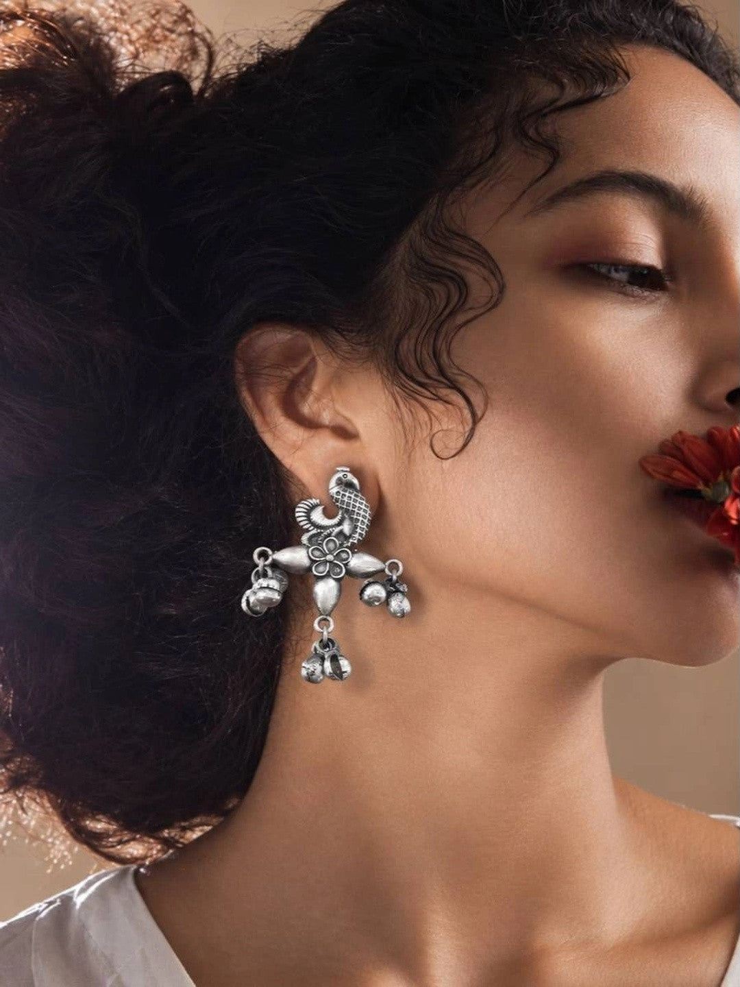 Oxidised silver peacock drop earring worn on a model's ear with red flower