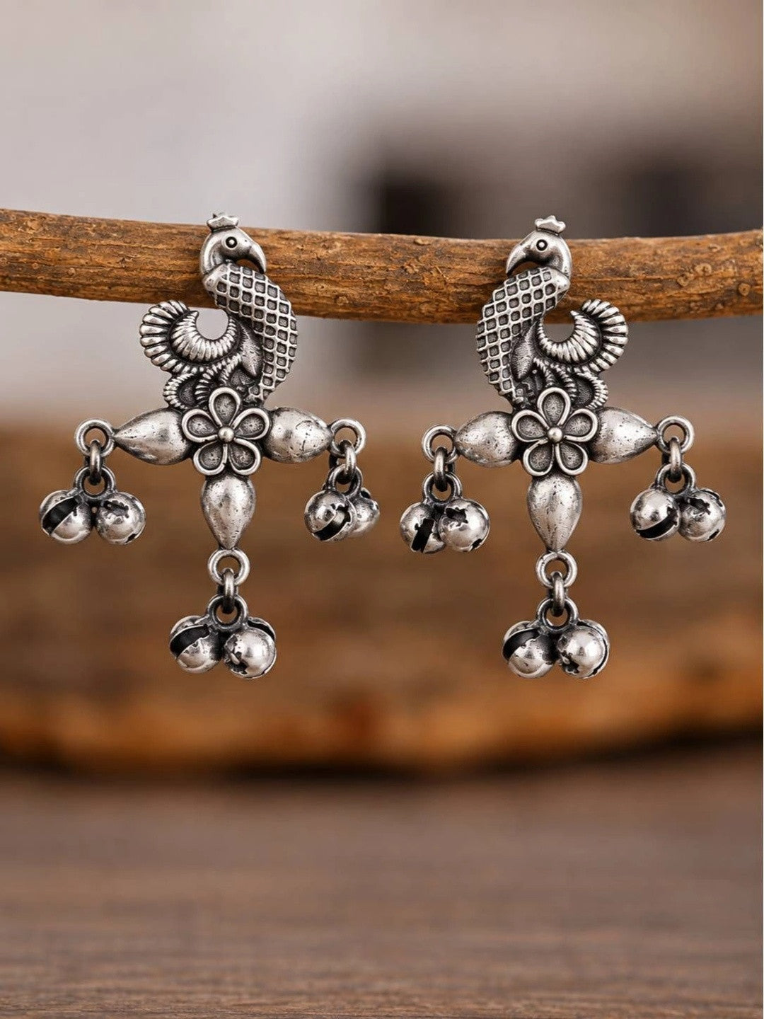 Close-up of two oxidised silver peacock earrings hanging from a wooden branch