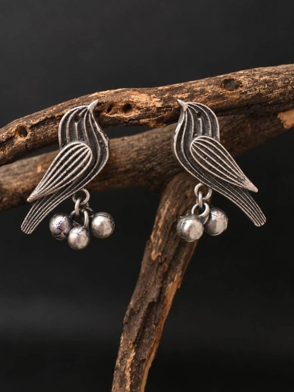 Two oxidised silver sparrow earrings with engraved feather detailing and dangling ghungroo bells displayed on a wooden branch