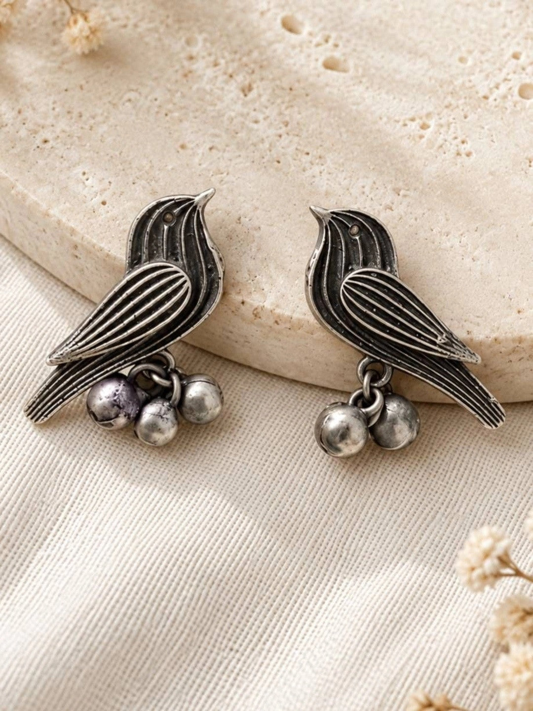 Pair of oxidised silver bird earrings with ghungroo drops resting against a textured stone prop, front view
