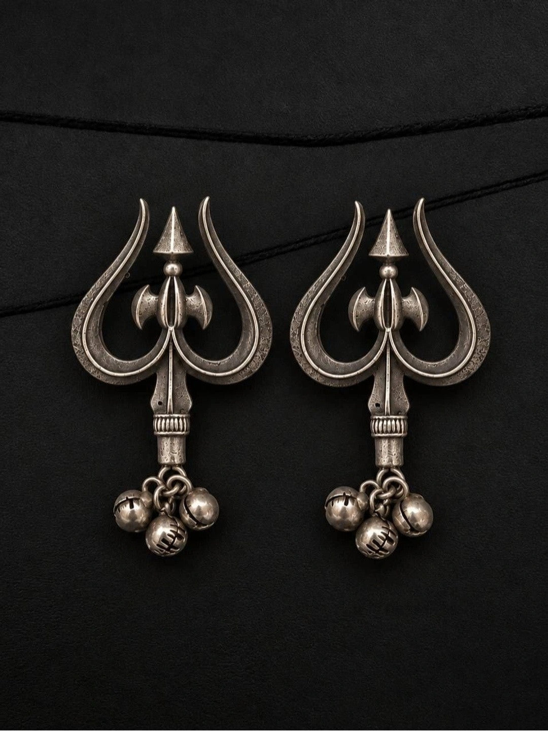 Pair of oxidised silver trishul drop earrings with parsu motif and three ghungroo bells laid flat on black surface