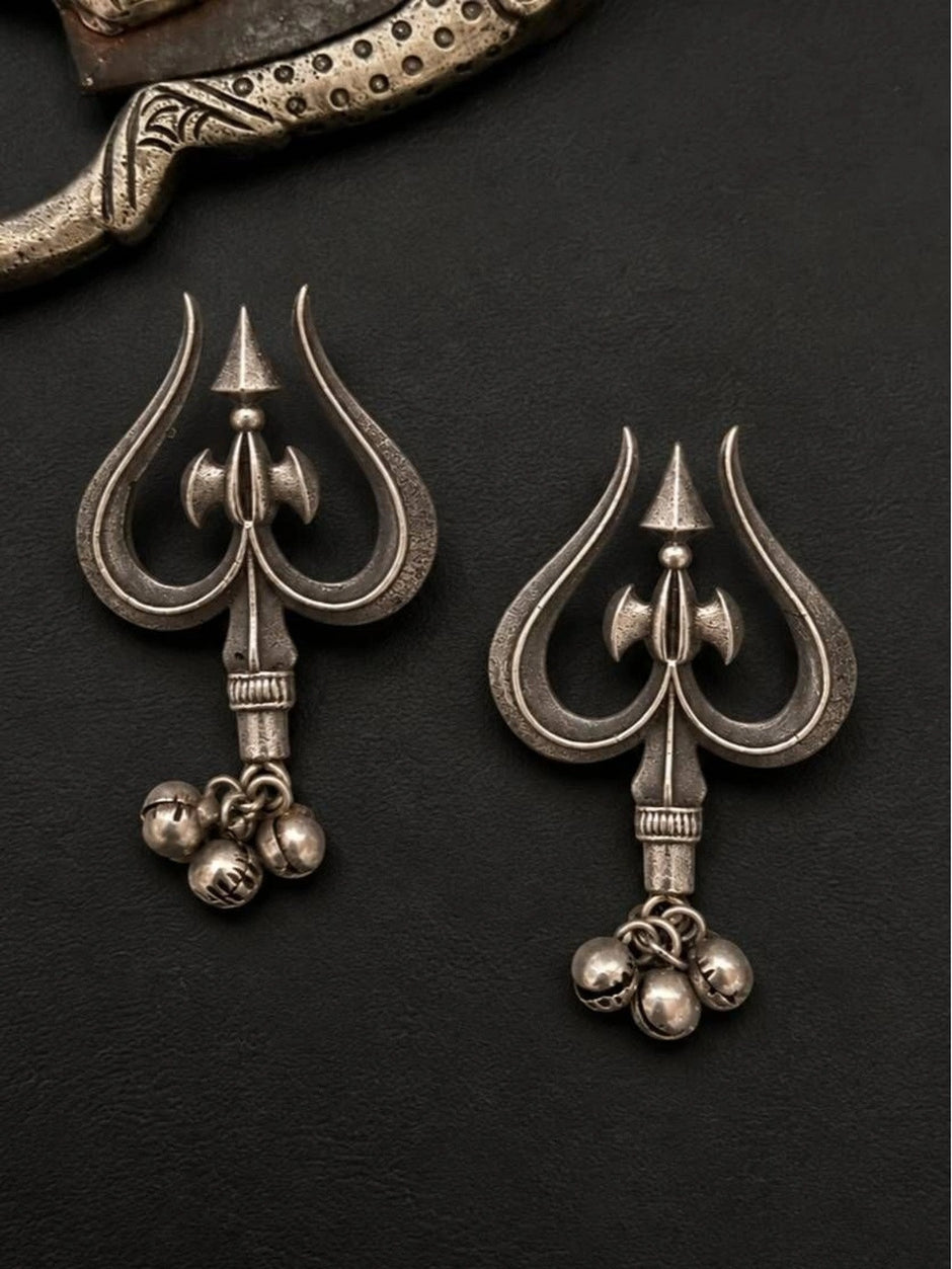 Close-up of two oxidised silver trishul earrings with antique finish and dangling ghungroo bells on dark background
