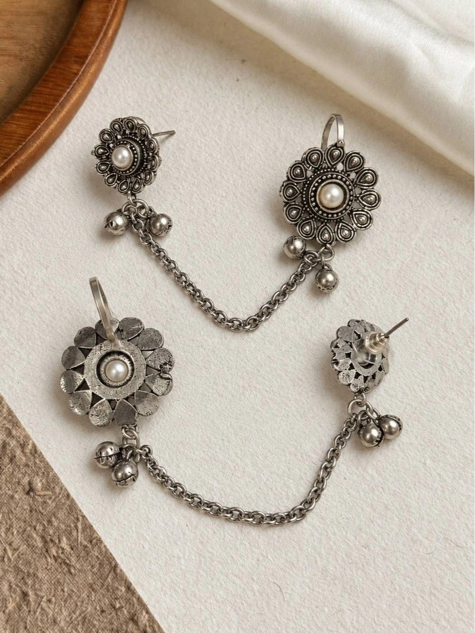 Two pairs of oxidised silver floral ear cuff studs with pearl centres, ghungroo drops, and linking chains laid flat on white surface