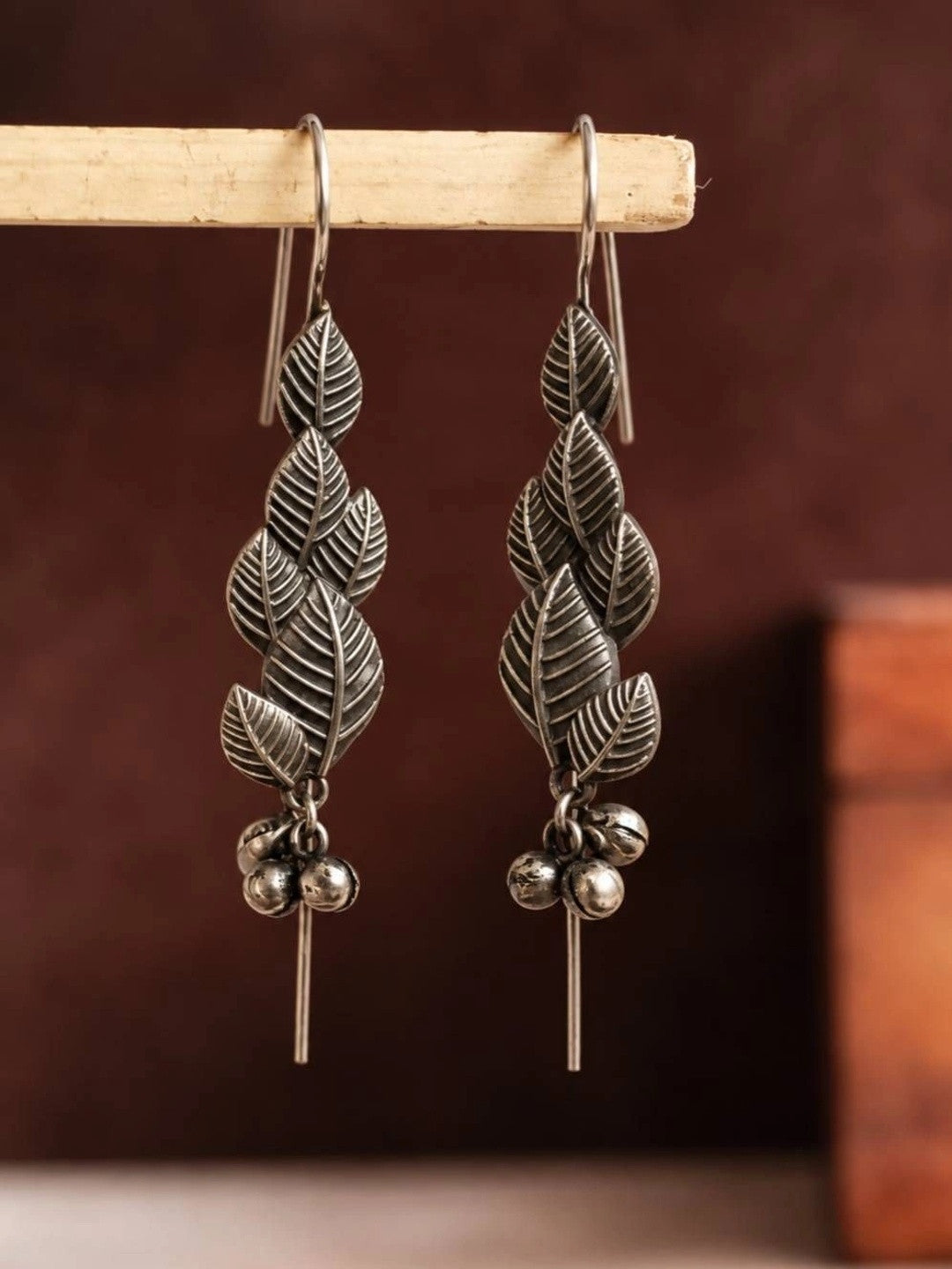 Pair of oxidised silver leaf drop earrings hanging on a wooden display rod with dark background
