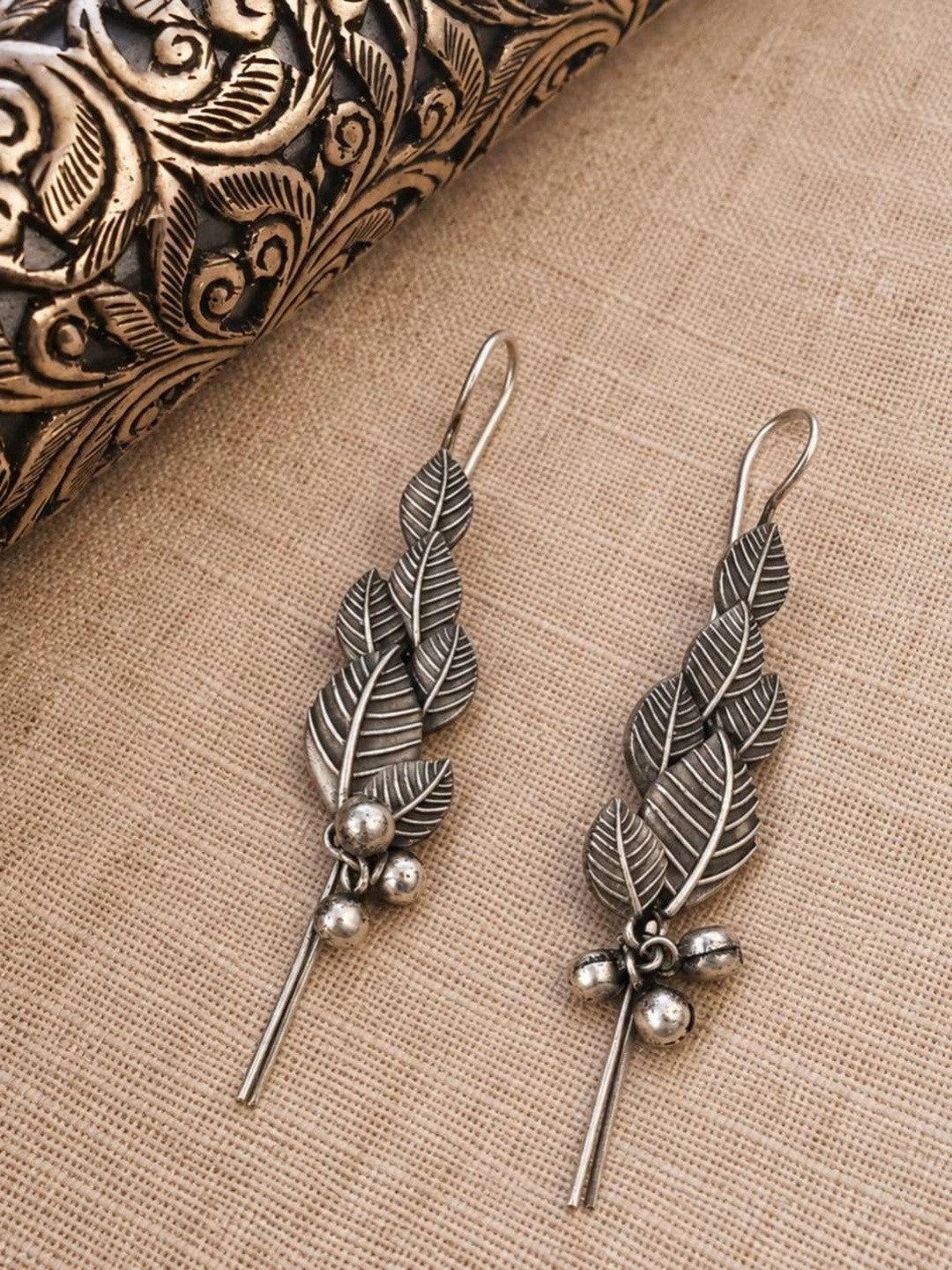 Pair of oxidised silver leaf earrings with ghungroo bells laid flat on linen beside a carved box