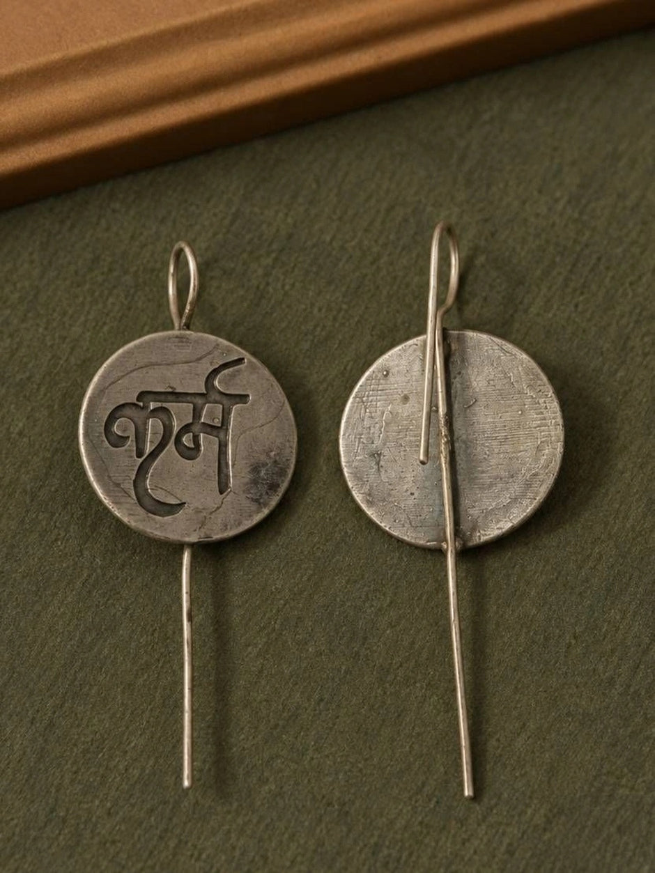 Front and back view of a single oxidised silver karma coin earring on a dark green surface showing the plain reverse side