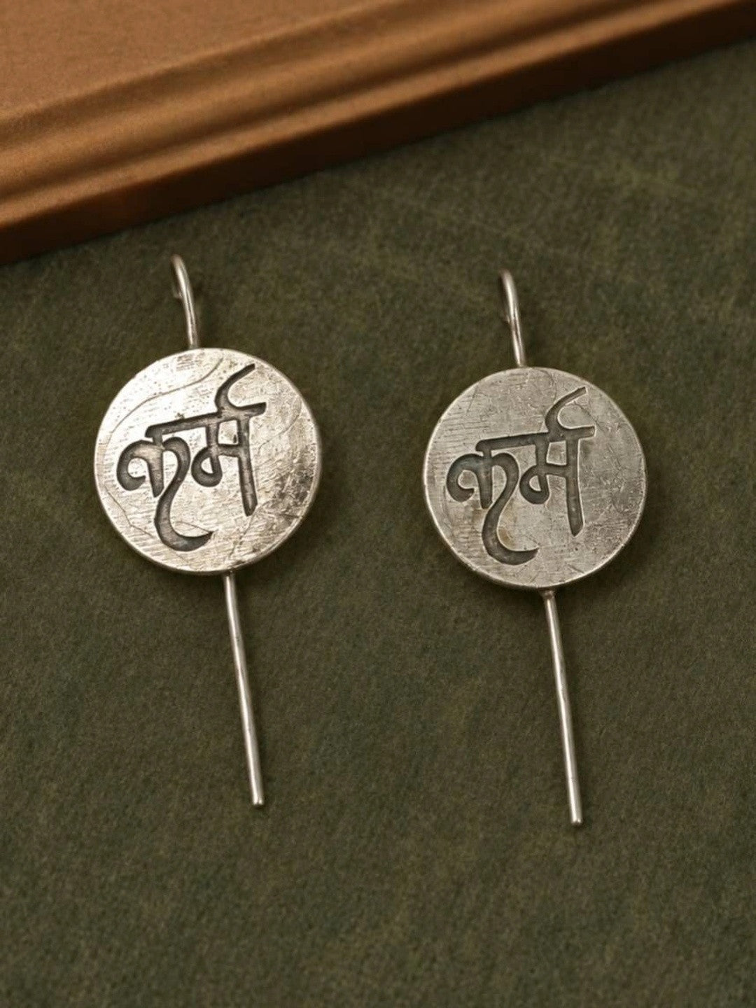 Two oxidised silver karma coin drop earrings laid flat on a dark green surface showing Devanagari engraving