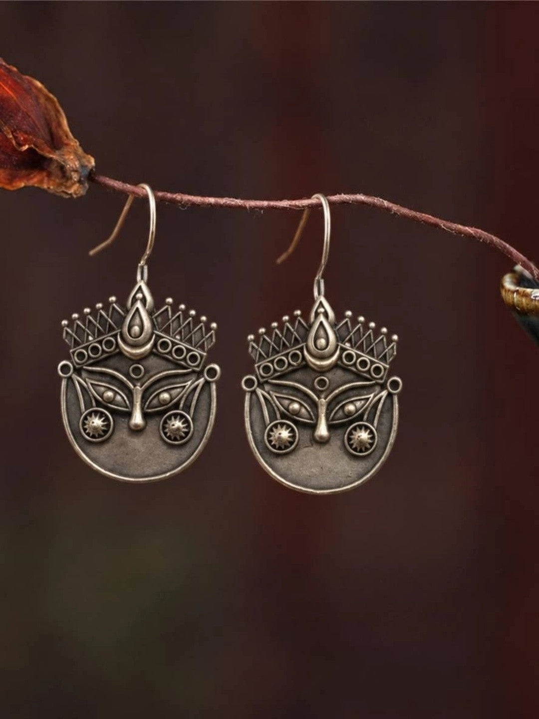 Pair of oxidised silver Durga face drop earrings hanging from a dried branch against a dark background