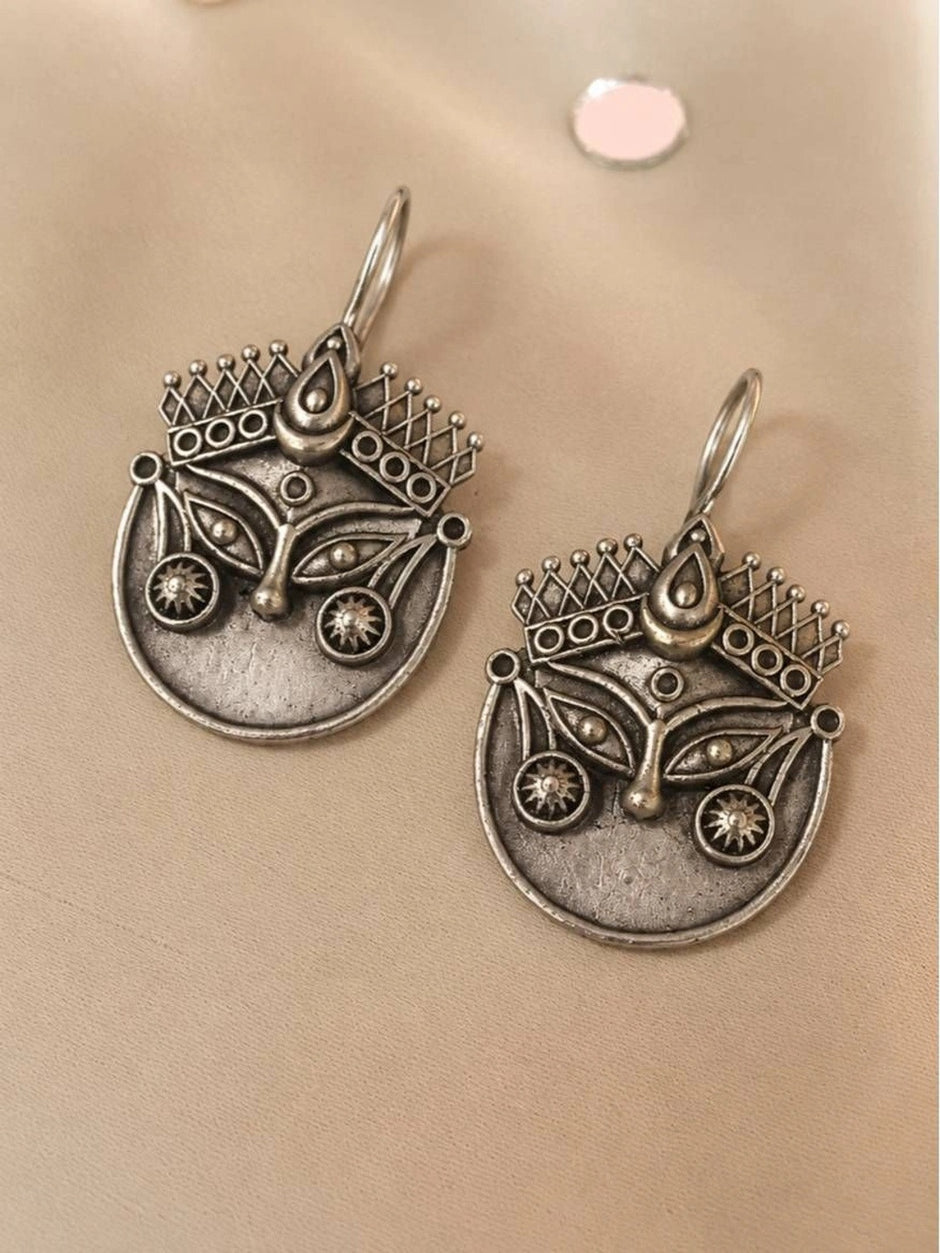Two oxidised silver Durga face earrings with crown and eye detailing laid flat on a cream surface