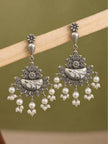 Two oxidised silver floral chandbali earrings with pearl drops hanging from a wooden display rod against green background