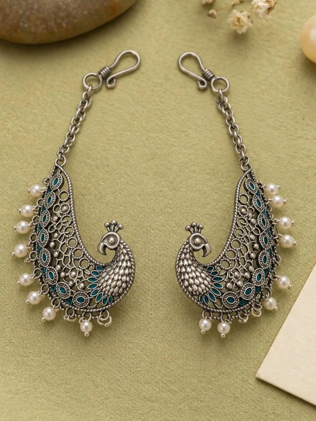 Pair of oxidised silver peacock ear cuff earrings with teal enamel and white pearl drops on sage green background