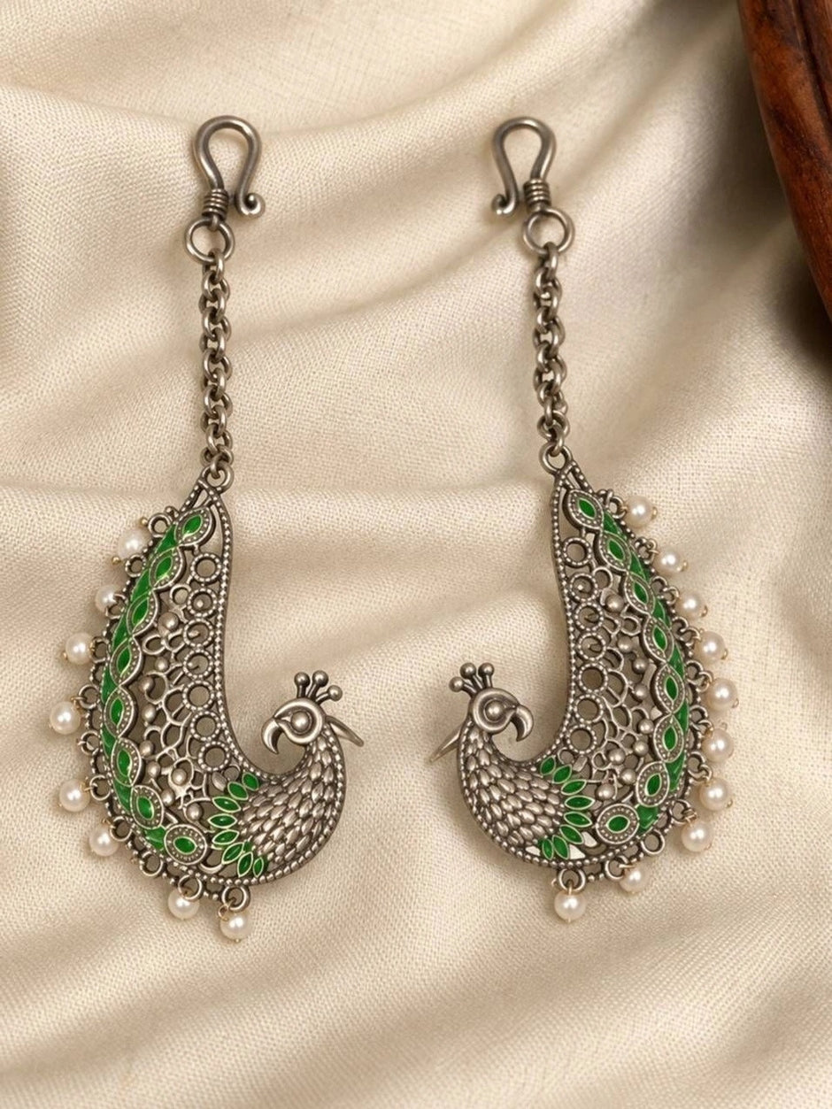 Pair of oxidised silver peacock ear cuffs with green enamel and white pearl drops laid flat on cream fabric