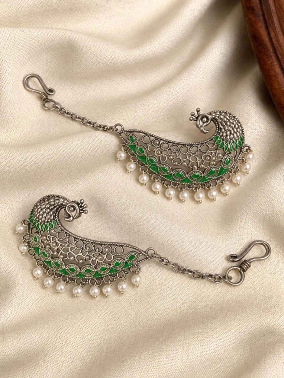 Two oxidised silver peacock ear cuffs facing opposite directions showing green enamel wings and dangling pearl beads