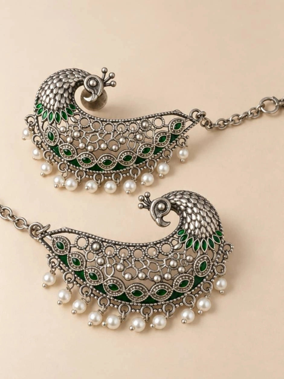 Close-up flat lay of two peacock ear cuff earrings showing intricate silver texture and pearl drop fringe