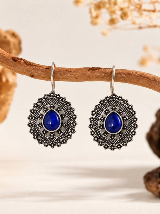 Pair of oxidised silver floral medallion drop earrings with blue cabochon stones hanging from a wooden branch