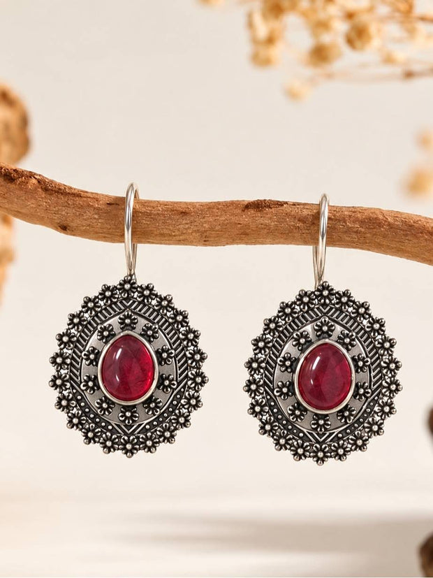 Pair of oxidised silver floral disc drop earrings with red cabochon stone hanging on a wooden branch