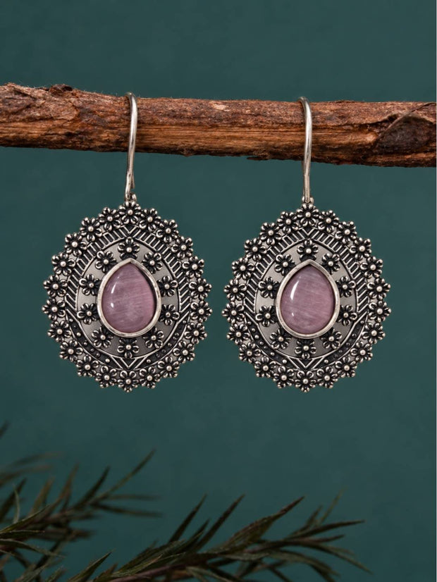 Pair of oxidised silver floral drop earrings with pink stone hanging from a wooden branch against teal background