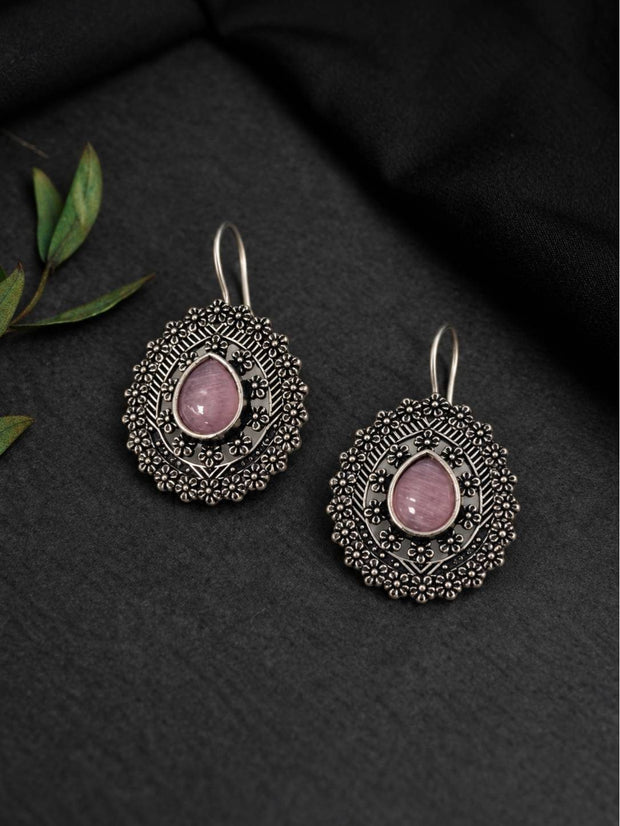Two oxidised silver mandala disc earrings with pink cabochon centres laid flat on dark fabric with green leaves