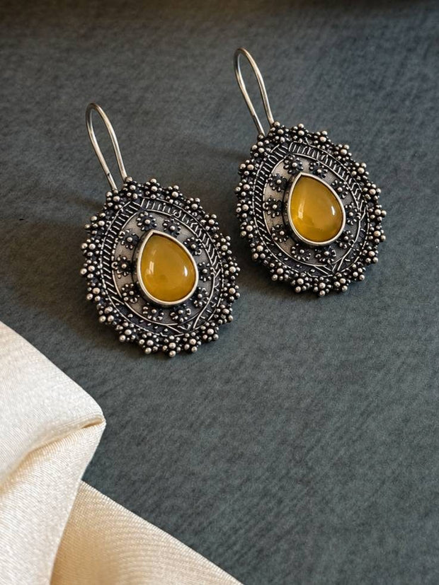 Two oxidised silver medallion earrings with yellow stone centres laid flat on a dark grey surface beside cream fabric