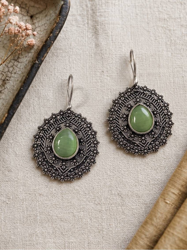 Oxidised silver drop earrings with green oval stones and concentric floral detailing laid flat on cream linen fabric