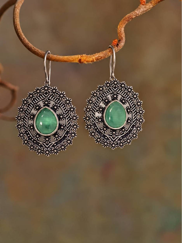 Pair of oxidised silver floral oval drop earrings with green teardrop cabochon hanging from a rustic branch