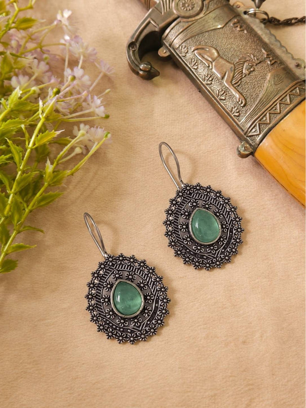 Two oxidised silver oval drop earrings with green stone centres laid flat on a beige surface beside purple flowers