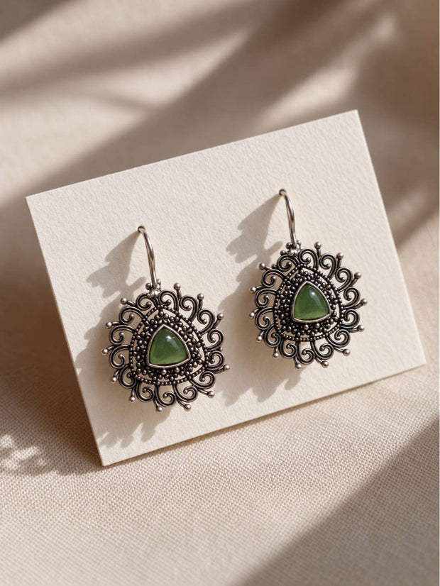 Pair of oxidised silver filigree drop earrings with green triangular stone displayed on a white card against a warm beige background