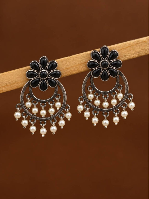 Pair of oxidised silver chandbali earrings with black floral tops and ivory pearl drops displayed on a wooden rod against a brown background