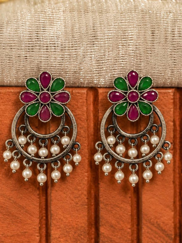 Pair of oxidised silver chandbali earrings with red and green floral stone tops and white pearl drop dangles displayed against a wooden surface