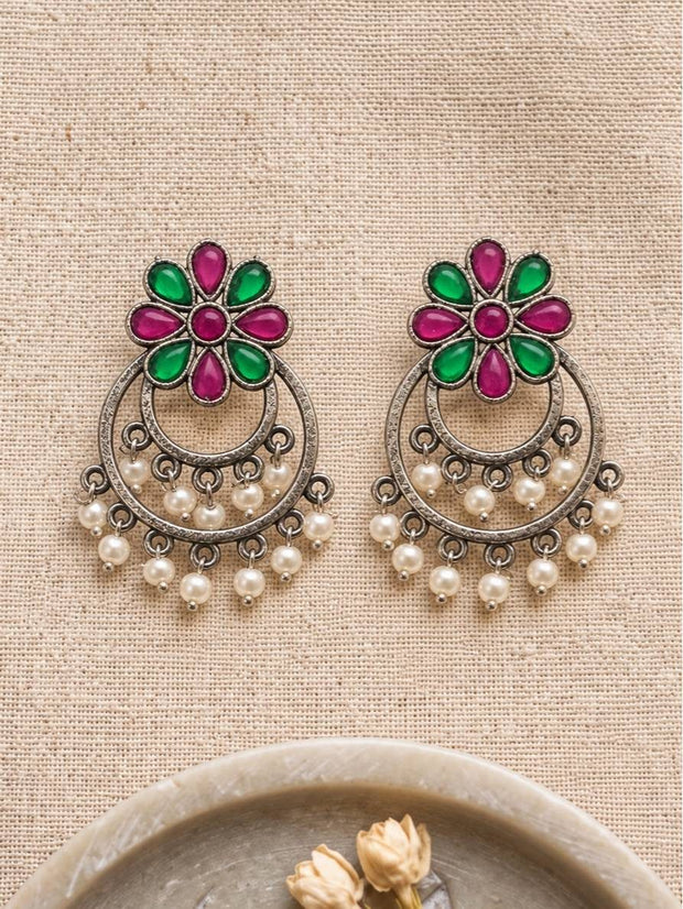 Front view of both oxidised silver chandbali earrings laid flat showing red green flower tops and pearl drops on a cream fabric background