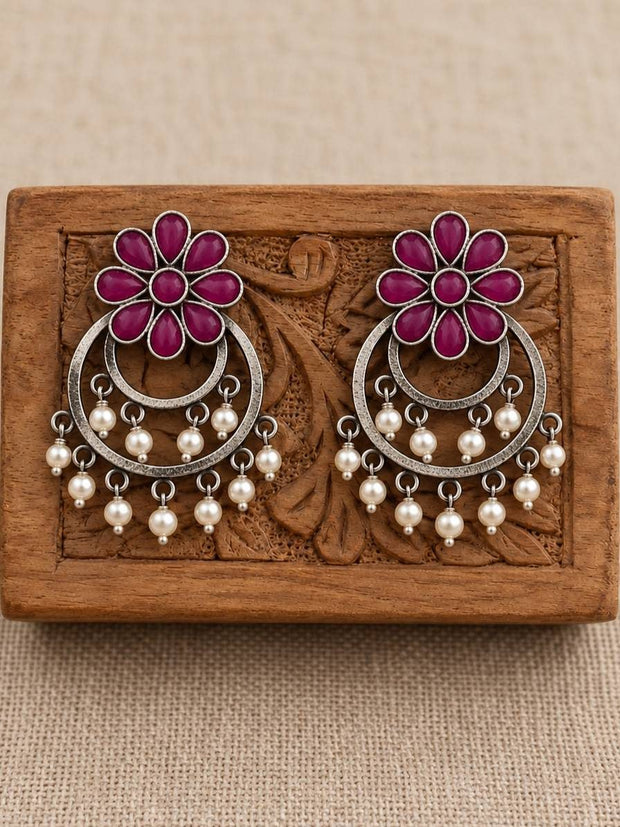 Pair of oxidised silver chandbali earrings with magenta floral tops and pearl drops displayed on a carved wooden block