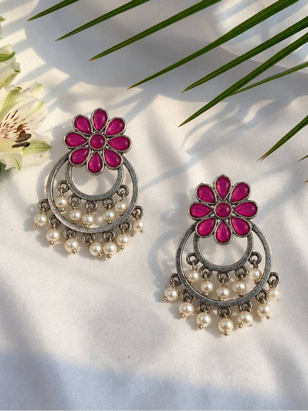 Front view of two oxidised silver chandbali earrings with magenta glass flower and pearl dangles on white fabric