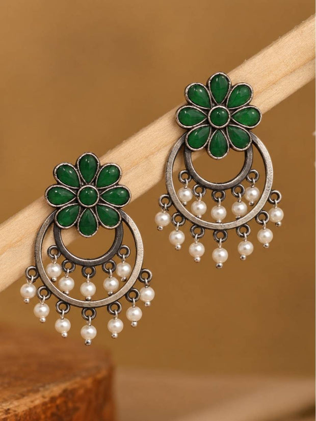 Pair of oxidised silver chandbali earrings with green flower stone tops and white pearl drops hanging on a wooden rod