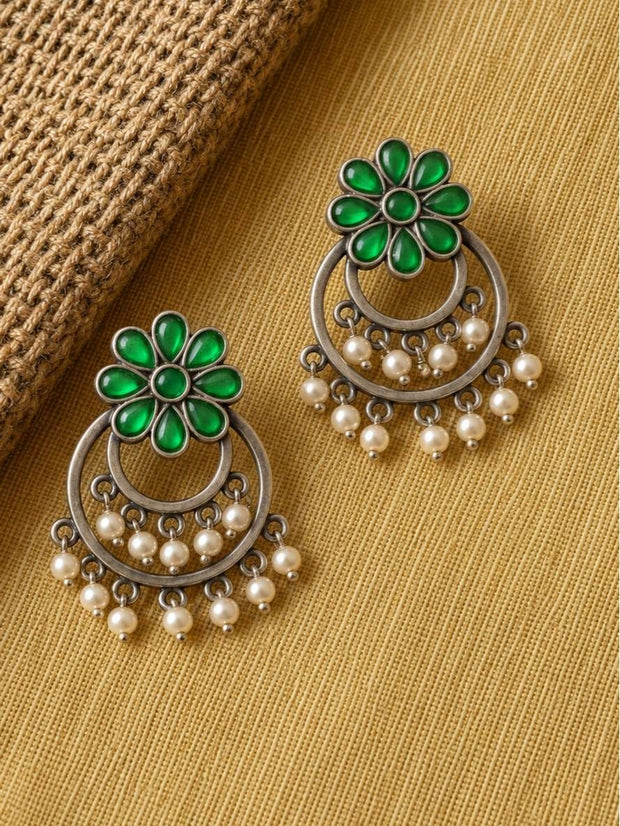 Two oxidised silver green stone chandbali earrings with pearl drops laid flat on a yellow woven textile surface