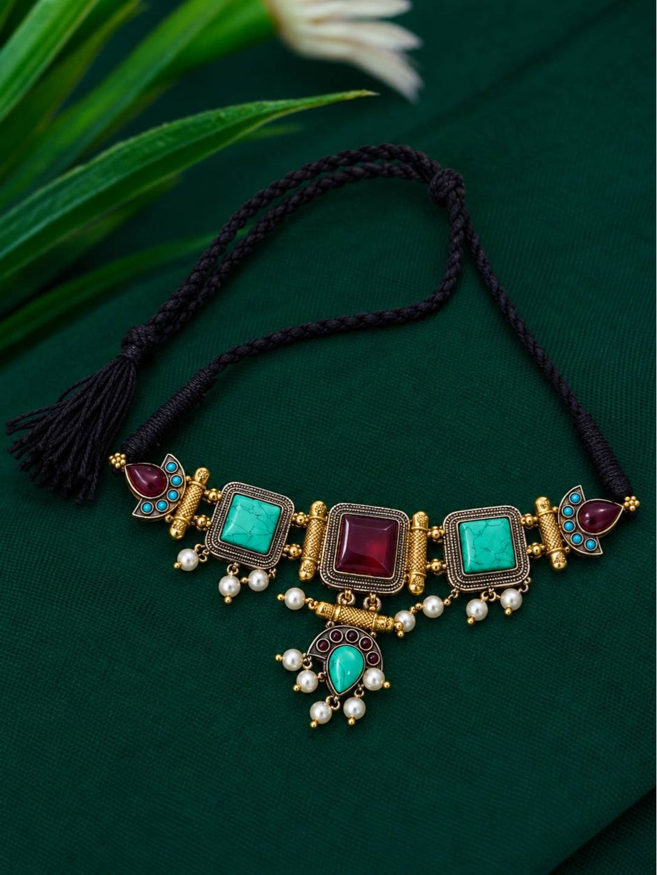 Antique gold turquoise and maroon stone choker necklace laid flat on dark green fabric with black dori cord and tassel