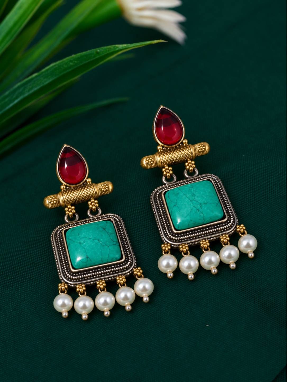 Close-up of a pair of antique gold turquoise square dangler earrings with maroon cabochon tops and pearl drops on green fabric