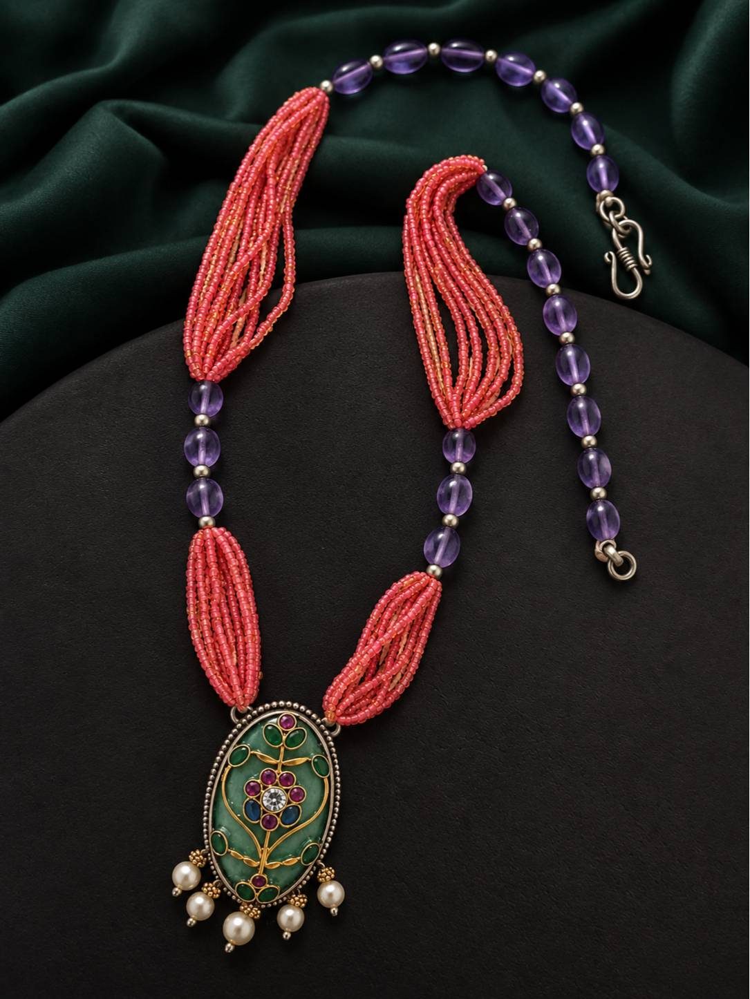 Coral seed bead and amethyst bead meenakari pendant necklace laid flat on dark green fabric showing S-hook clasp