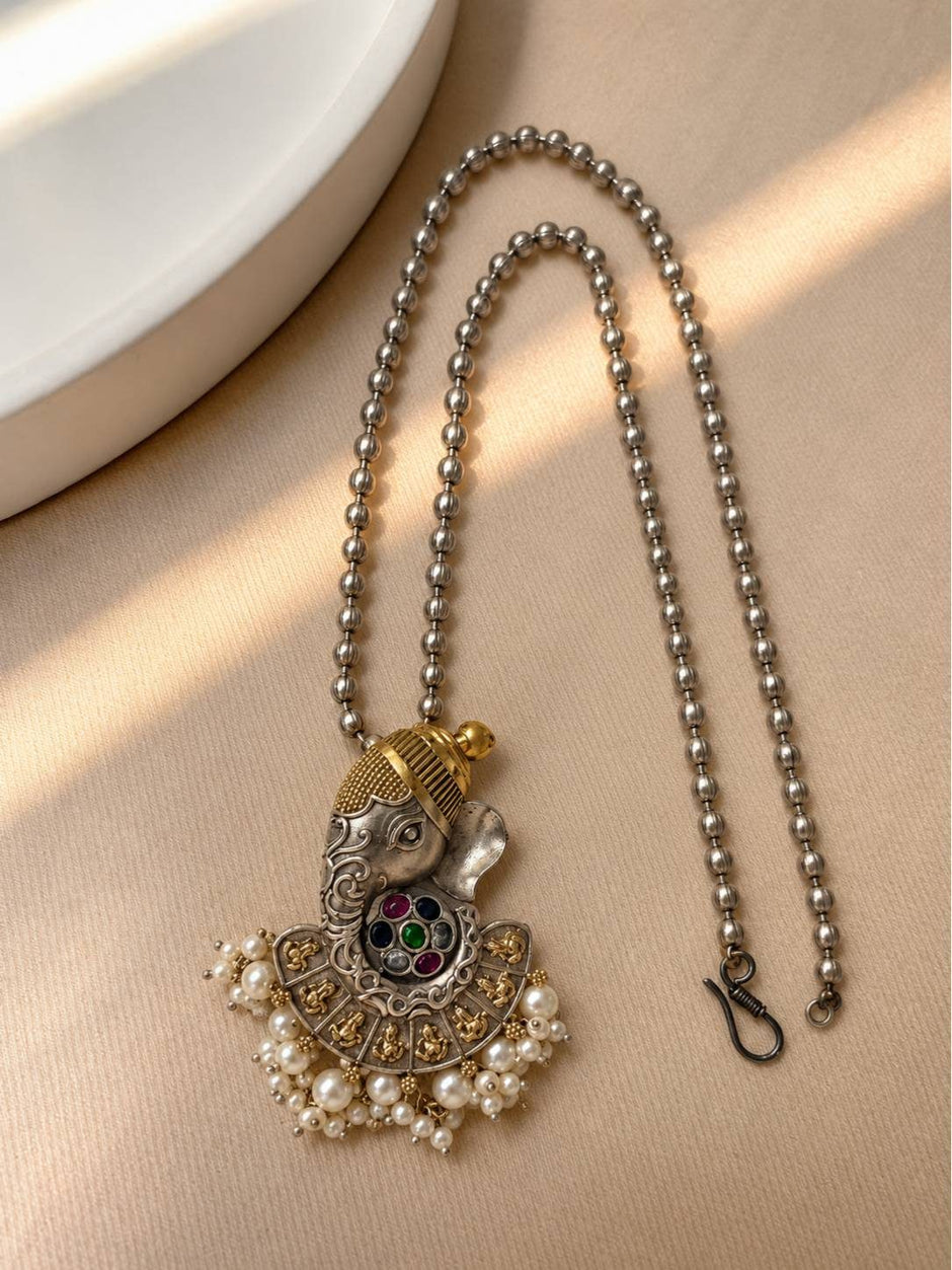 Close-up of Ganesha pendant on oxidised silver ball-chain mala laid flat on a beige surface showing S-hook clasp