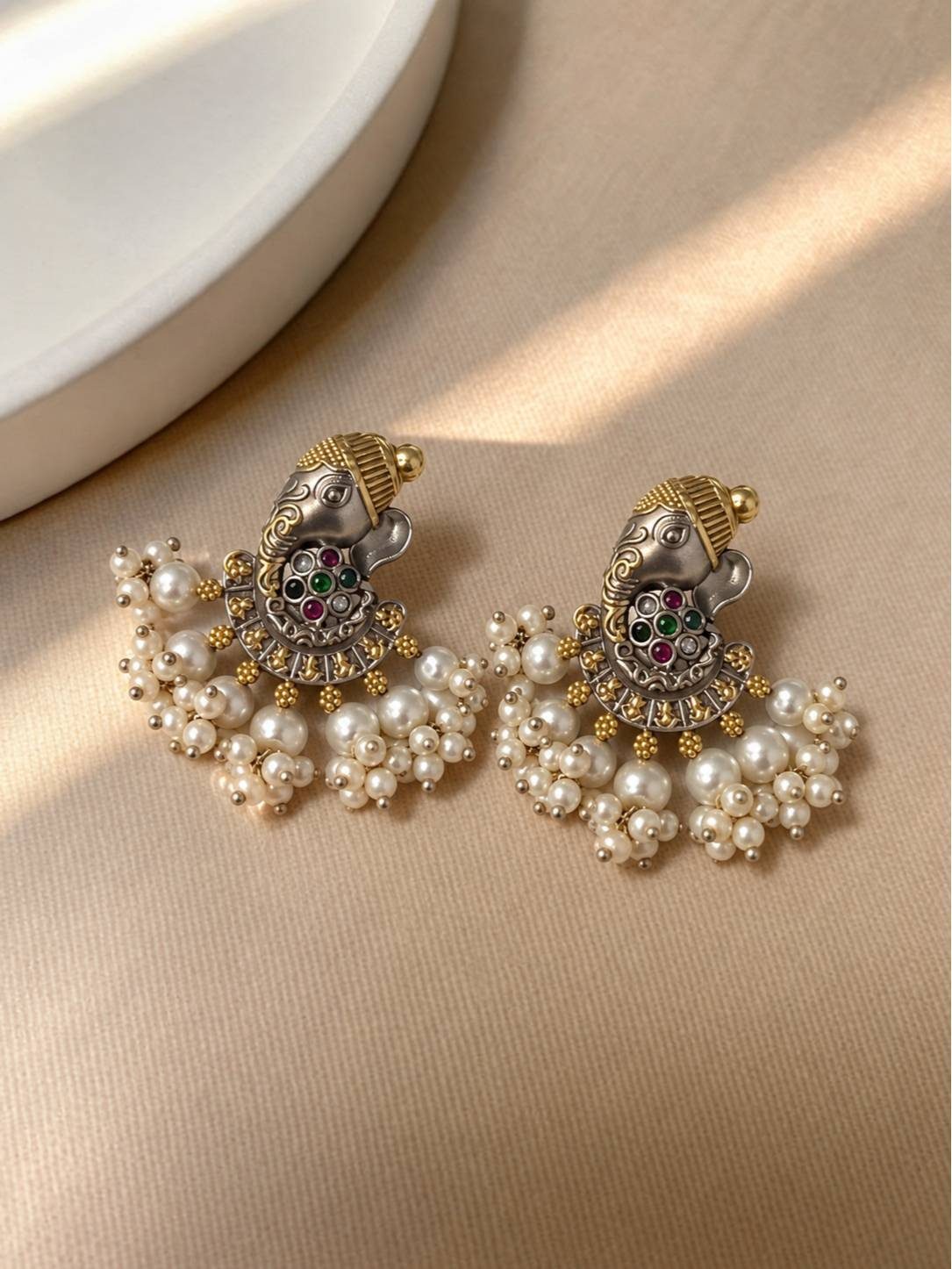 Pair of dual-tone Ganesha chandbali earrings with multicolour stones and faux pearl drops on a beige surface